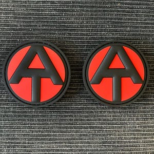 以下が含まれることがあります： 「A」と「T」の文字が書かれた黒と赤の丸いパッチが2つ。パッチは灰色のテクスチャのある表面に置かれています。