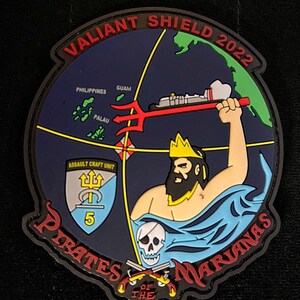 May include: A black and red embroidered patch with a blue background. The patch features a map of the Pacific Ocean with the text "Valiant Shield 2022" around the top. The patch also features a trident, a skull and crossbones, and the text "Pirates of the Marianas" around the bottom.