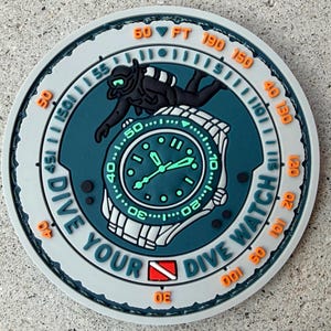 May include: A circular rubber patch featuring a teal and gray design with a diver silhouette and a wristwatch graphic. The text DIVE YOUR DIVE WATCH is embossed in gray, with orange depth markers in feet ranging from 30 to 190, and a red and white diver down flag.