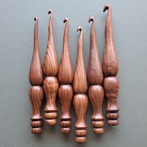 Op de afbeelding: Set van zes houten haaknaalden met bruine en roodbruine houtnerf. De haaknaalden hebben allemaal dezelfde vorm, met een afgeronde handgreep en een puntige haak.