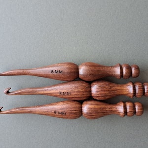 Op de afbeelding: Drie houten haaknaalden, elk met een maat van 9,0 mm gemarkeerd op de handgreep. De haaknaalden hebben een donkerbruine houtnerf en een afgeronde handgreep met een knop aan het einde.