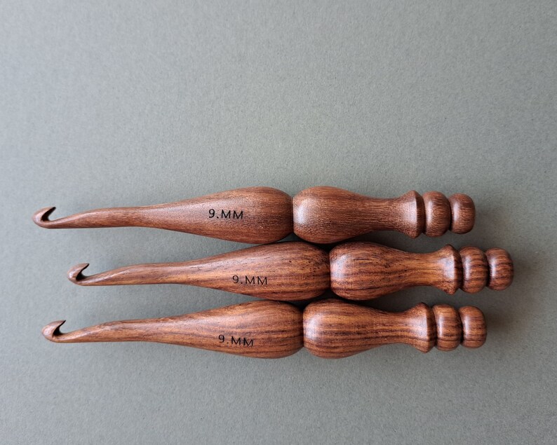 Op de afbeelding: Drie houten haaknaalden, elk met een maat van 9,0 mm gemarkeerd op de handgreep. De haaknaalden hebben een donkerbruine houtnerf en een afgeronde handgreep met een knop aan het einde.