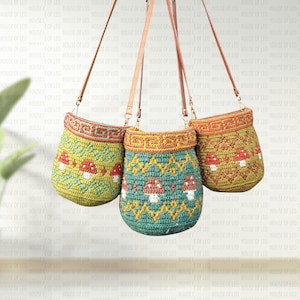 Op de afbeelding: Drie handgemaakte gehaakte schoudertassen in groen en blauwgroen, versierd met paddenstoelmotieven. Elke tas heeft een bruine leren riem en een decoratief geometrisch patroon aan de bovenkant. De tassen hangen tegen een witte achtergrond.