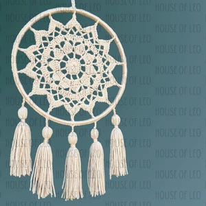 May include: A white crocheted dreamcatcher with a circular design and five tassels hanging from the bottom. The dreamcatcher is hanging against a teal background.