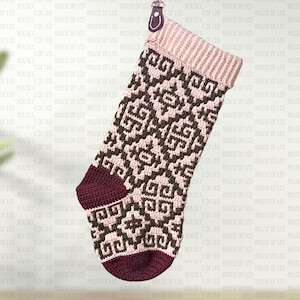 Pode incluir: Uma meia de Natal em crochê com um punho canelado rosa claro e um padrão geométrico repetitivo em castanho escuro e rosa claro. O calcanhar e a biqueira são de uma cor bordô sólida, e uma pequena alça de couro está presa no topo.