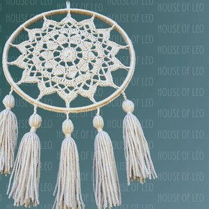 May include: A white crocheted dreamcatcher with a circular frame and a starburst design. The dreamcatcher has five white tassels hanging from the bottom.
