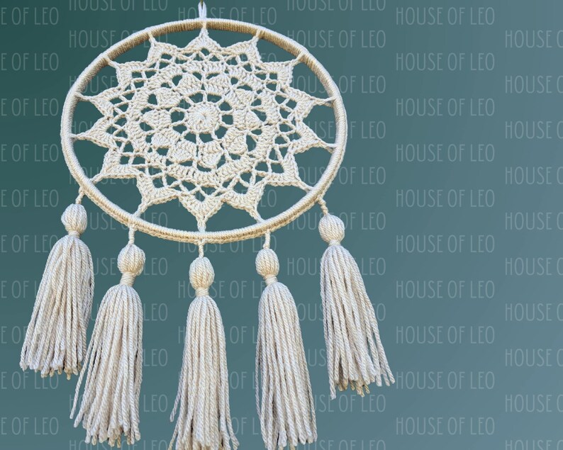 May include: A white crocheted dreamcatcher with a circular frame and a starburst design. The dreamcatcher has five white tassels hanging from the bottom.