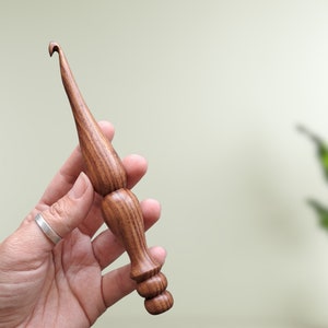 Op de afbeelding: Een houten haaknaald met een bruin en zwart houtnerfpatroon. De haaknaald wordt vastgehouden in een hand met een zilveren ring.