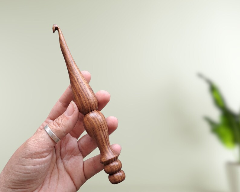 Op de afbeelding: Een houten haaknaald met een bruin en zwart houtnerfpatroon. De haaknaald wordt vastgehouden in een hand met een zilveren ring.