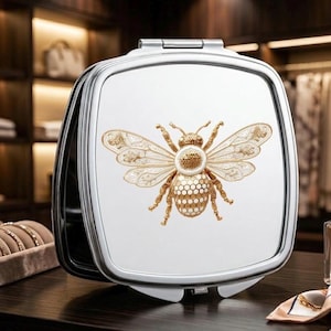 May include: A silver compact mirror with a square shape and a hinged opening. The mirror's surface features a detailed gold-colored bee design with intricate wing patterns and a honeycomb-patterned body. The mirror is open, revealing the reflective surface.