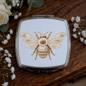 May include: A square, silver compact mirror with a detailed gold-colored bee design on a light blue background. The bee has intricate wing and body patterns. The mirror is surrounded by white flowers and greenery.