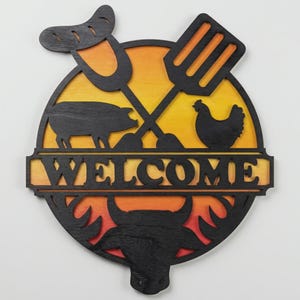 May include: A circular wooden sign with a black outline and a yellow and orange gradient background. It features crossed grilling tools, a pig, a chicken, a sausage, and a bull's head. The word "WELCOME" is cut out at the bottom.