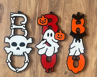 May include: Three Halloween-themed wooden signs spelling "BOO". The "B" is made of bones and a skull, the "O" has a ghost and pumpkins, and the other "O" has a bat and ghost. The signs are white, black, red, and orange.