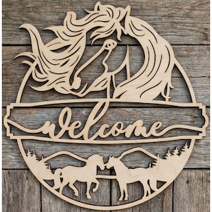 Puede incluir: Letrero de bienvenida de madera con diseño de caballos. El letrero incluye la palabra "welcome" en cursiva, dos caballos, montañas y árboles. Hecho de madera clara, adecuado para uso en interiores o exteriores. Forma circular.