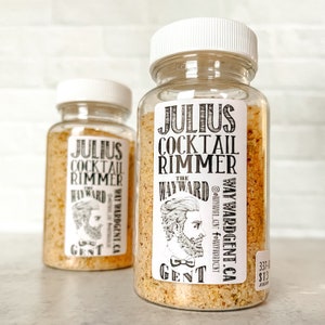 May include: Two clear plastic jars filled with a light brown granular substance, labeled "Julius Cocktail Rimmer" with a vintage illustration of a man. The jars have white screw-on lids and are set against a white brick wall.