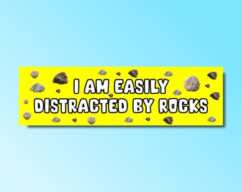 May include: A yellow banner with the text "I am easily distracted by rocks" in white letters. The banner is decorated with small gray and brown rocks.