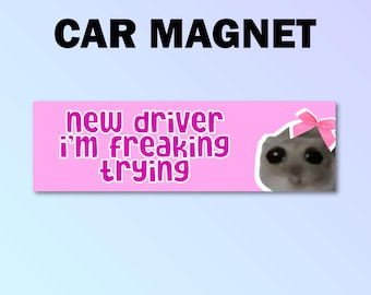 以下が含まれることがあります： ピンクの車のマグネット。白い猫がピンクのリボンをつけており、「new driver i'm freaking trying」というピンクの文字が書かれています。