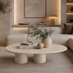 May include: An oval-shaped beige travertine coffee table with two cylindrical pedestal bases. The table surface holds two stacked books, a small shallow bowl, and two rounded vases, one of which contains a leafy green branch.