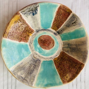 May include: A handmade ceramic plate with a unique design. The plate features a circular shape with segments of turquoise, brown, grey, and yellow. The centre has a smaller circle with a brown speckled pattern. The plate is a decorative piece.