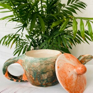 Puede incluir: Una tetera de cerámica hecha a mano con tapa. El cuerpo de la tetera es principalmente verde y naranja, con un patrón texturizado similar a burbujas. La tapa es naranja con un pomo redondeado. Objeto decorativo.