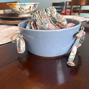 May include: A light blue ceramic bowl with a speckled finish, featuring a fabric liner with a floral pattern in shades of pink, green, and blue. The liner has a drawstring closure with fabric ties that extend over the sides of the bowl.
