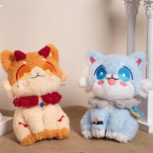 以下が含まれることがあります： 2つのぬいぐるみ猫のおもちゃ。1つはオレンジとクリーム色で赤いアクセントがあり、もう1つは水色と白です。どちらも大きな目を持ち、直立しています。オレンジ色の猫は赤い角と赤い首輪をしています。青い猫は灰色の首輪をしています。