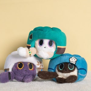 Könnte beinhalten: Drei Plüschtiere in Form von Tierköpfen, die Helme tragen. Das erste Spielzeug ist ein weißer Plüsch mit einem grünen Helm mit einem schwarzen Pik darauf. Das zweite Spielzeug ist ein lilafarbener Plüsch mit einem weißen Helm mit goldenen Akzenten. Das dritte Spielzeug ist ein brauner Plüsch mit einem blauen Helm mit einer weißen Wolke darauf.