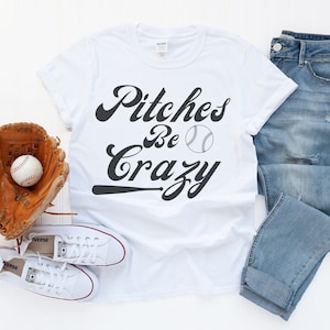 May include: White t-shirt with a black graphic that says "Pitches Be Crazy" with a baseball and a baseball bat. The shirt is laid out on a white surface with a baseball glove, a baseball, and a pair of white sneakers.