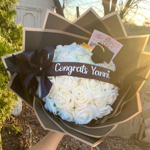 May include: A bouquet of white roses with pearl accents, wrapped in dark green and black paper. A black ribbon with the text "Congrats Yanni" is tied around the flowers. A small graduation cap and card are attached.
