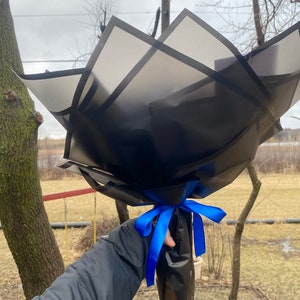 Puede incluir: Un ramo de flores envuelto en papel negro con una cinta azul. El papel tiene un patr&oacute;n geom&eacute;trico de cuadrados negros.