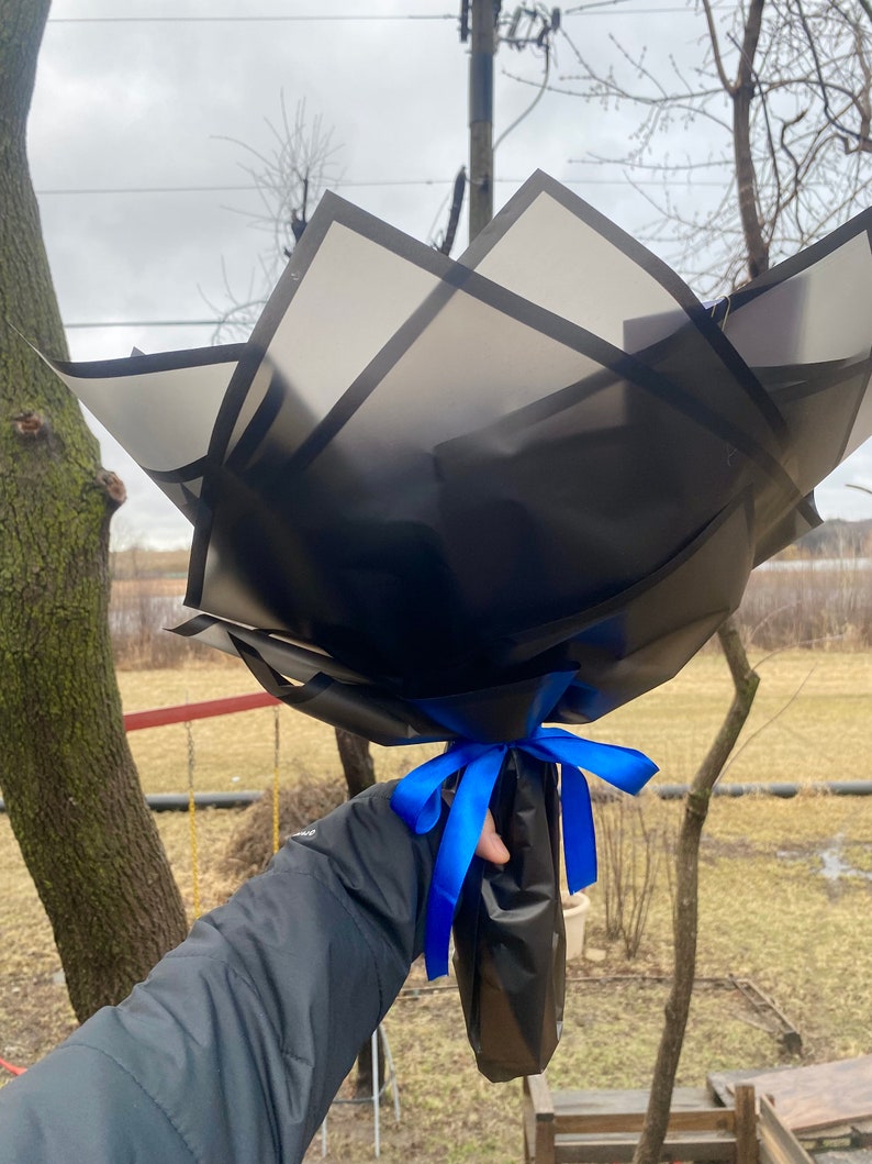 Puede incluir: Un ramo de flores envuelto en papel negro con una cinta azul. El papel tiene un patr&oacute;n geom&eacute;trico de cuadrados negros.