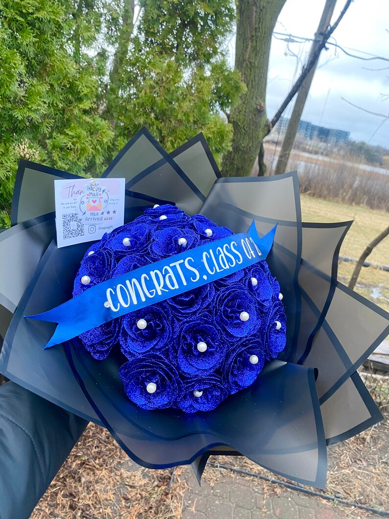 Puede incluir: Un ramo de graduaci&oacute;n azul y blanco con una cinta azul que dice "&iexcl;Felicidades, Clase de!" El ramo est&aacute; envuelto en papel de seda negro.