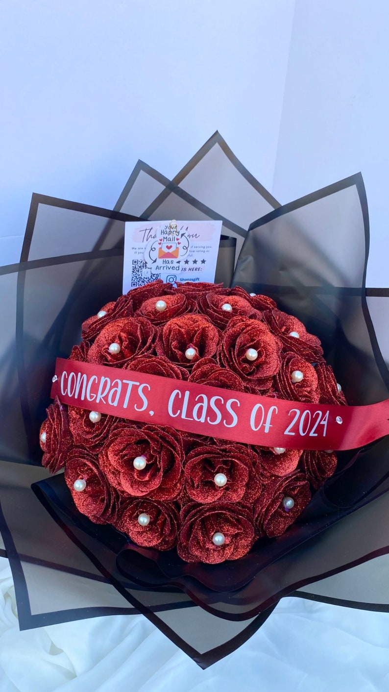 Puede incluir: Un ramo de rosas rojas con purpurina y detalles de perlas blancas, atado con una cinta roja que dice "Felicidades, Clase de 2024".