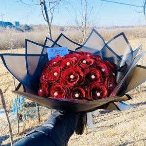 Puede incluir: Un ramo de rosas rojas de fieltro con detalles de perlas, envuelto en papel blanco y negro con una tarjeta que dice "The Heart of You".