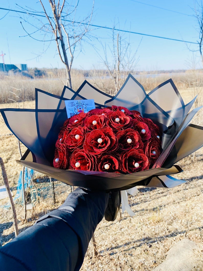 Puede incluir: Un ramo de rosas rojas de fieltro con detalles de perlas, envuelto en papel blanco y negro con una tarjeta que dice "The Heart of You".