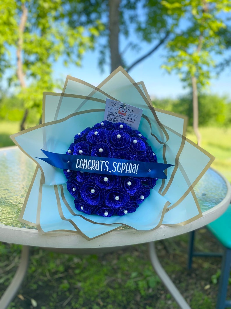 Puede incluir: Un ramo de flores de papel azul con detalles de perlas blancas. El ramo est&aacute; envuelto en papel azul claro y dorado y tiene una cinta azul con el texto "&iexcl;Felicidades, Sophia!"