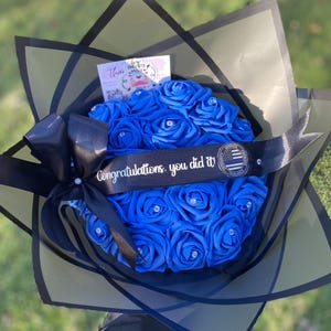 May include: A bouquet of vibrant blue artificial roses, each featuring a small clear rhinestone at its center. A black satin ribbon is wrapped around the flowers, printed with the text "Congratulations, you did it!" and a circular police emblem.