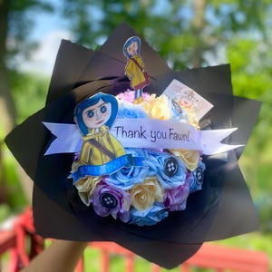 Puede incluir: Un ramo de flores artificiales de color pastel dispuestas en un envoltorio de papel negro. El ramo incluye una cinta blanca con el texto "&iexcl;Gracias Fawn!" y dos recortes de papel de personajes de la pel&iacute;cula "Coraline".
