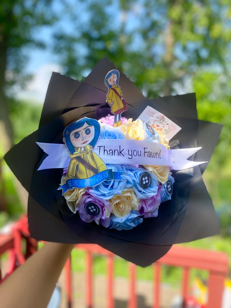 Puede incluir: Un ramo de flores artificiales de color pastel dispuestas en un envoltorio de papel negro. El ramo incluye una cinta blanca con el texto "&iexcl;Gracias Fawn!" y dos recortes de papel de personajes de la pel&iacute;cula "Coraline".