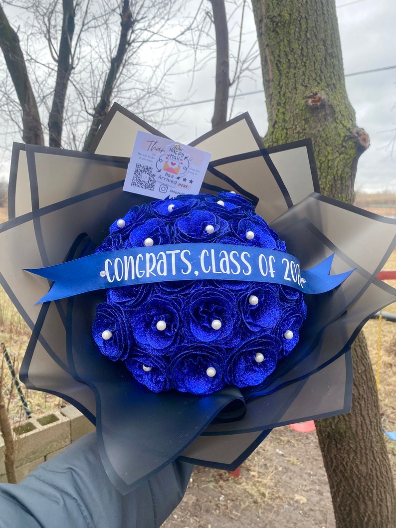 Puede incluir: Un ramo de rosas de terciopelo azul con detalles de perlas blancas. El ramo est&aacute; envuelto en papel de seda negro y una cinta azul con el texto "Felicidades, Clase de 202."