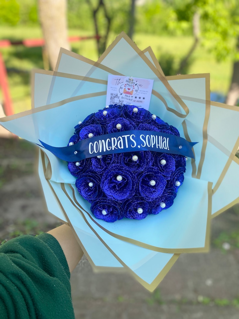 Puede incluir: Un ramo de flores artificiales azul y blanco con una cinta azul que dice "&iexcl;Felicidades, Sophia!" y una tarjeta peque&ntilde;a que dice "The Happy Mail".