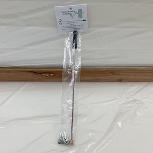 May include: A silver tufting needle with a black plastic handle, packaged in a clear plastic bag. The bag has a label that reads "Tufting Needle".