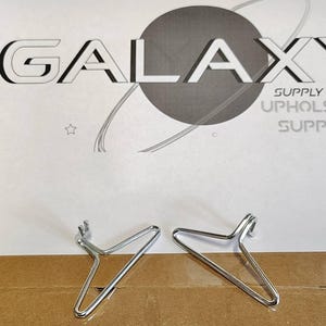 May include: Two silver metal upholstery tacks on a brown cardboard surface. The tacks are shaped like a Y and have a pointed end. The background is a white sheet of paper with the text "GALAXY SUPPLY INC. UPHOLSTERY SUPPLIES".