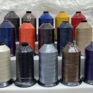 May include: A collection of spools of thread in various colors, including beige, black, brown, blue, gray, red, and orange. The spools are arranged in rows on a white surface.