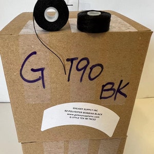 May include: Two spools of black thread, labeled "G190 BK", are stacked on top of a cardboard box. The box is labeled "Galaxy Supply Inc. 92 Polyester Bobbins Black www.galaxysupplyinc.com G Style Tex 90 Trcez".