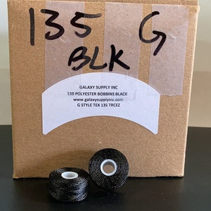 May include: A cardboard box labeled "135 G BLK" with two black thread spools in front. The box has a label that reads "GALAXY SUPPLY INC 138 POLYESTER BOBBINS BLACK". The spools are black with white centers.
