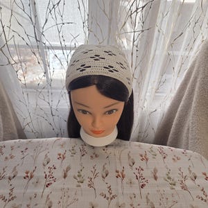 May include: A cream-colored crocheted head covering with a decorative openwork pattern. The head covering is displayed on a mannequin head with dark hair. The background includes a floral patterned fabric and a sheer curtain.
