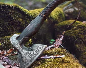 May include: A detailed close-up of a Viking axe with a dark wooden handle and a silver-colored head. The handle is wrapped in dark brown leather. The axe head features intricate engraved designs and symbols. The axe is resting on a mossy, natural surface.