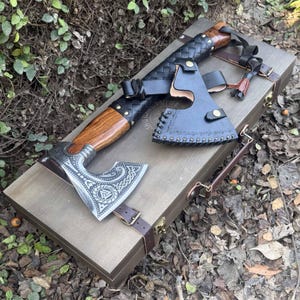 Könnte beinhalten: Eine handgefertigte Axt mit Holzgriff und dekorativem Metallkopf, auf einer Holzkiste liegend. Der Axtkopf weist aufwendige eingravierte Designs auf. Eine schwarze Lederscheide und Riemen sind enthalten. Die Kiste hat Lederriemen und Metallverschlüsse.