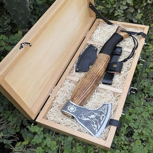 Puede incluir: Hacha con mango de madera y cabeza metálica decorativa, presentada en una caja de madera con paja. La cabeza del hacha presenta un diseño de sol y montaña. Incluye funda de cuero y trabilla para cinturón.